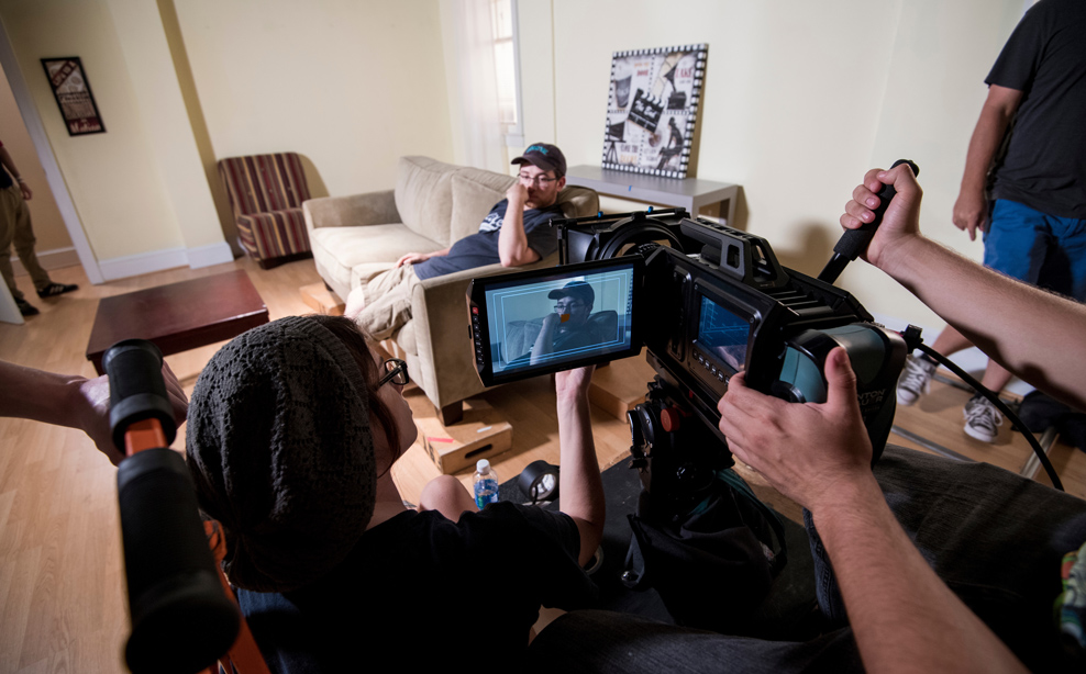 Students work a camera while another student sits on a couch in the frame