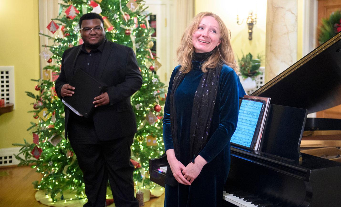 A woman stands at a piano with a singer in the background