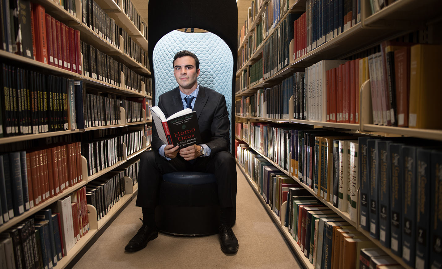 Man sitting in the Stacks at UNCW Library holding a copy of  Homo Deus