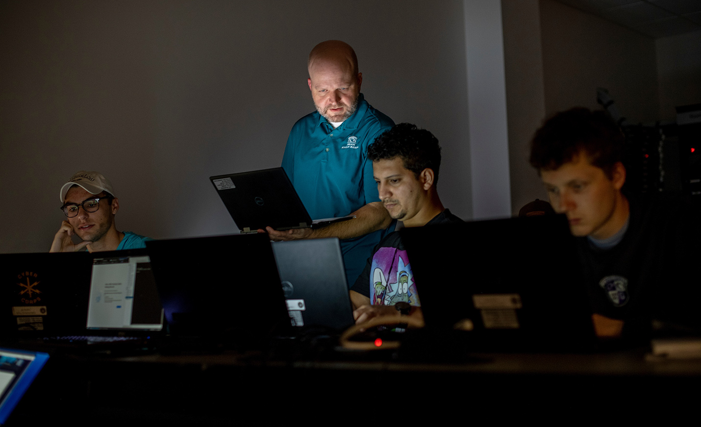 An instructor holding a laptop stands near two students who are working on laptops