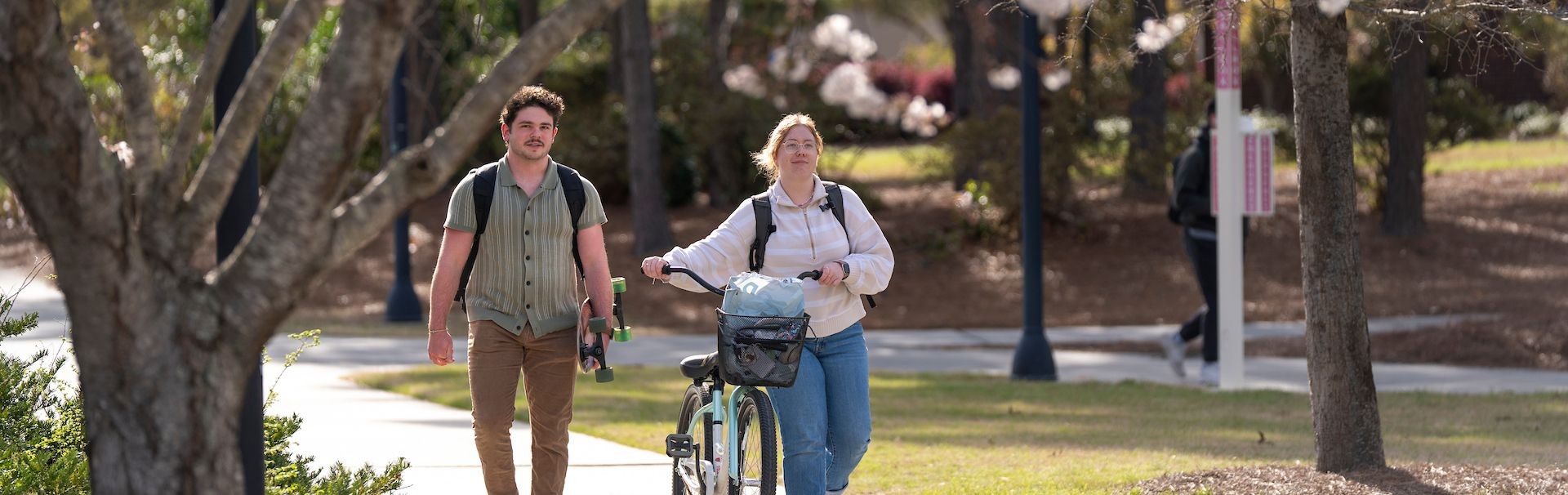 Two students walking on a sidewalk while one of them is wheeling a bike alongside