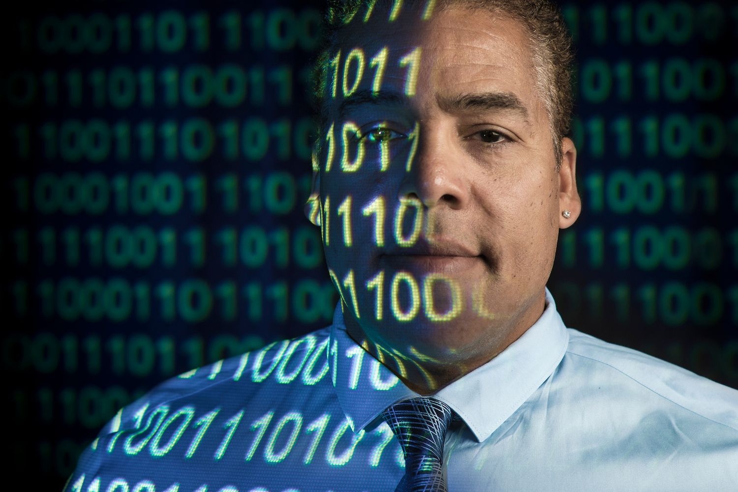 A professor looks at the camera with 1’s and 0’s projected onto his face 