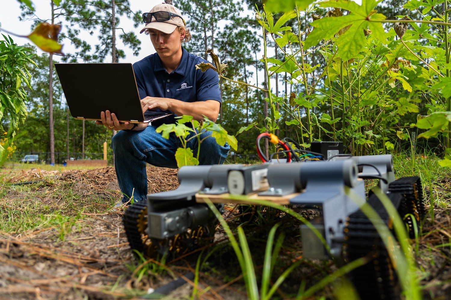 Person kneeling outdoors with a laptop, monitoring or controlling a small tracked robot equipped with sensors and wiring among green plants in a natural setting.