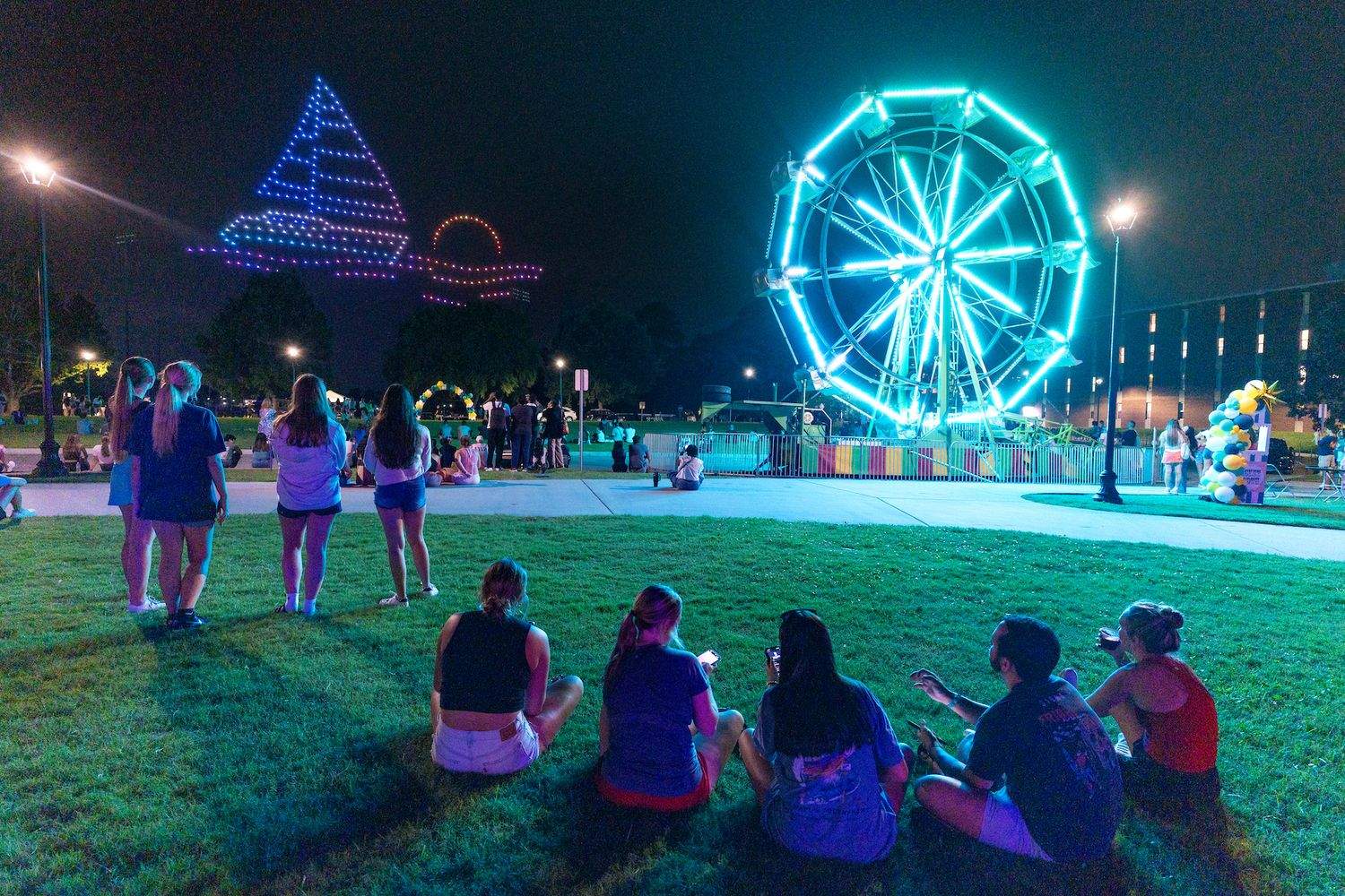 People watch a drone light show with carnival rides lit up in the night.